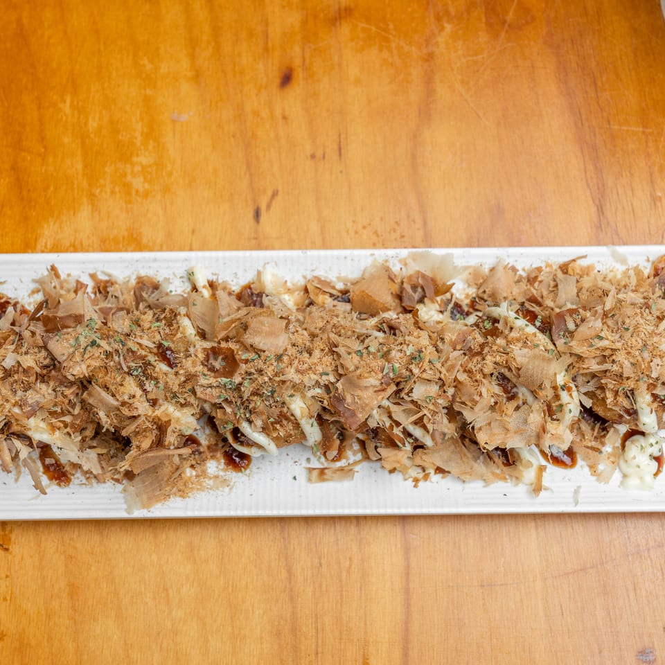 Long rectangular platter of shredded savory Japanese-style food topped with bonito flakes, sauces, mayonnaise, and herbs on a wooden table.