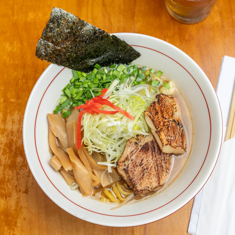 Bowl of ramen topped with grilled pork, bamboo shoots, shredded cabbage, scallions, red peppers, and seaweed on a wooden table.