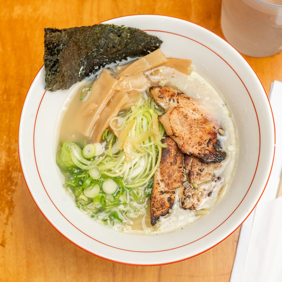Bowl of Japanese ramen with grilled chicken, bamboo shoots, scallions, noodles, and seaweed on a wooden table.