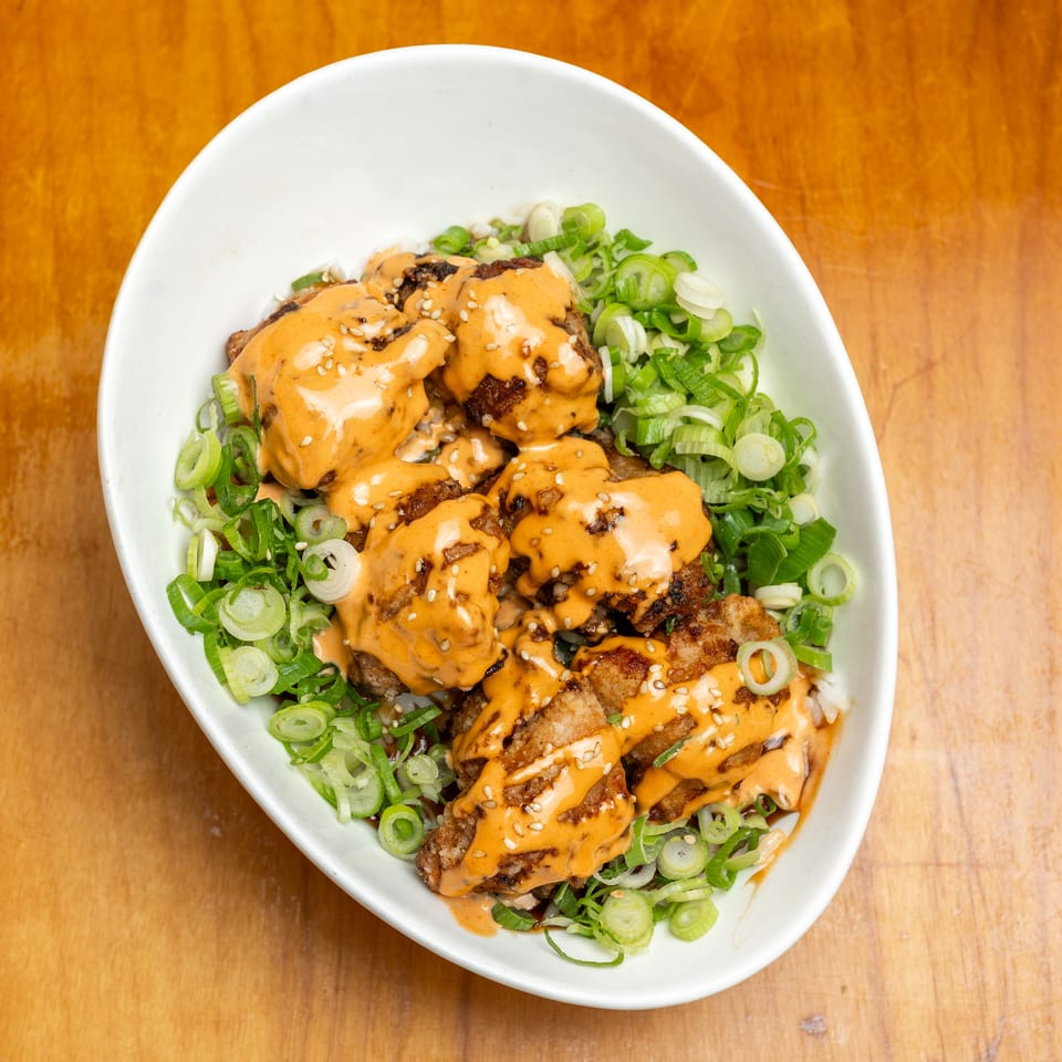 Bowl of rice topped with crispy chicken, orange sauce, sesame seeds, and sliced green onions on a wooden table.