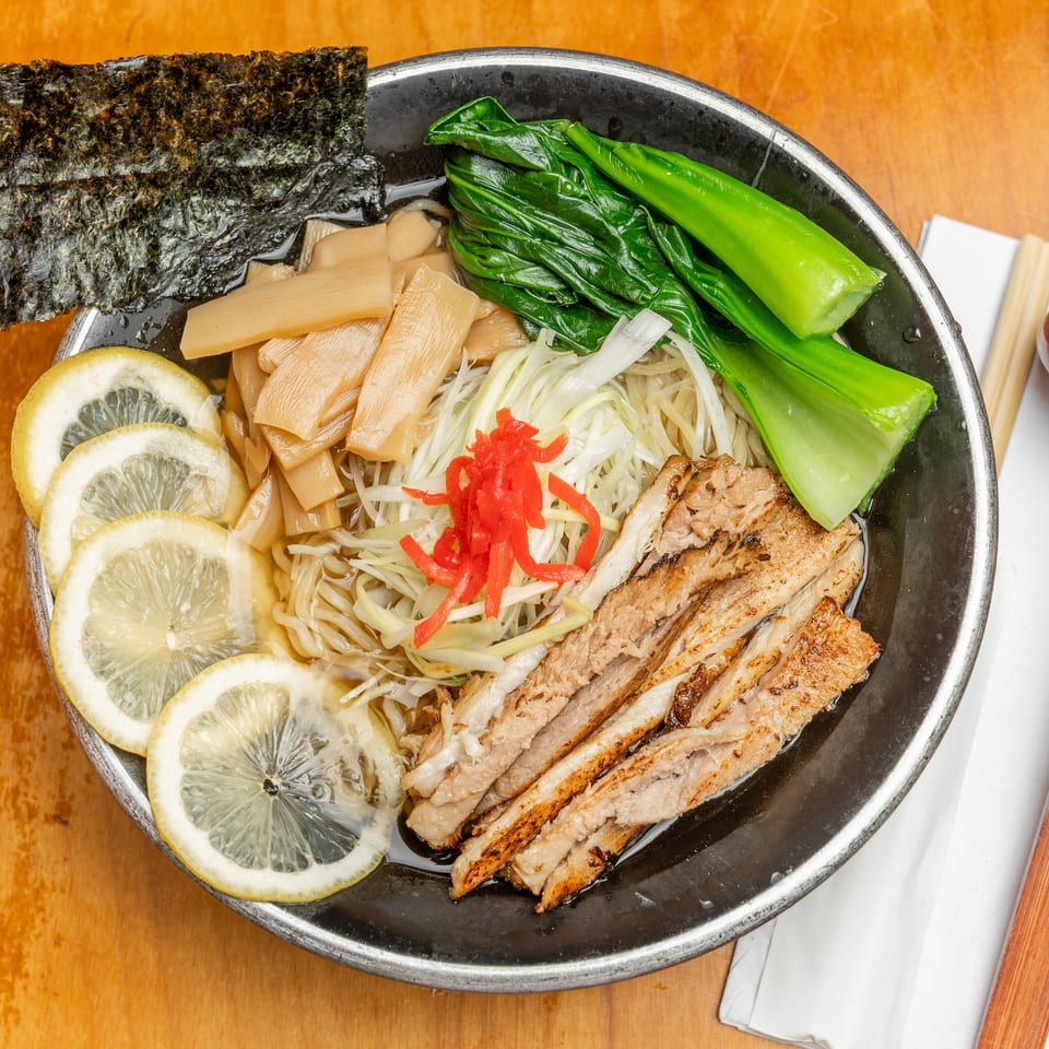 Japanese ramen bowl topped with grilled chicken, greens, bamboo shoots, lemon slices, shredded vegetables, red ginger, and seaweed.