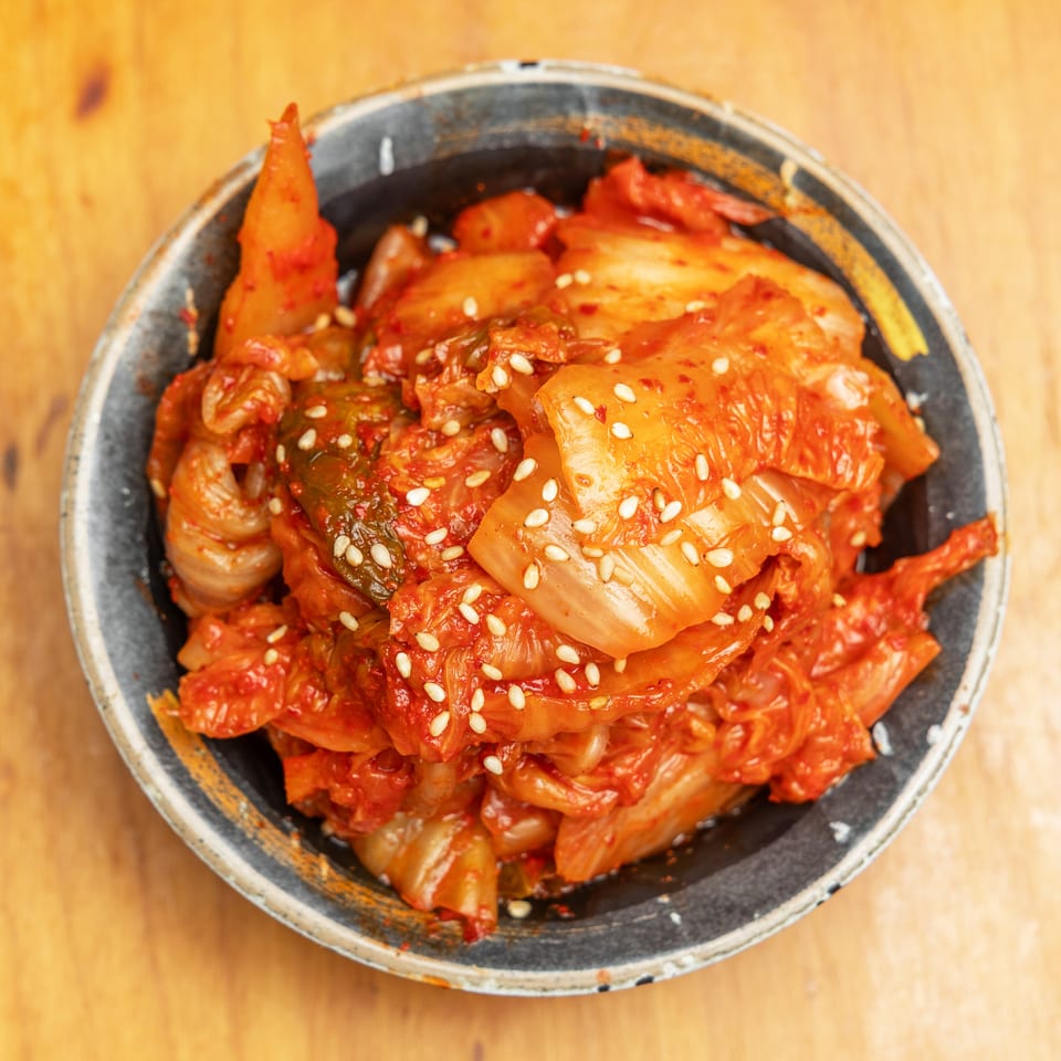 Spicy red kimchi topped with sesame seeds in a rustic bowl on a wooden table.