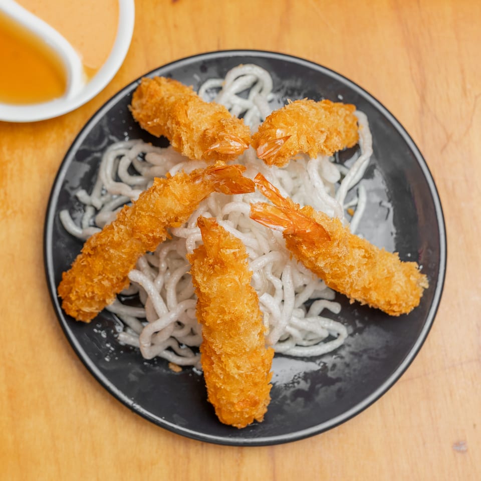 Crispy breaded shrimp served over noodles on a black plate with orange dipping sauce.