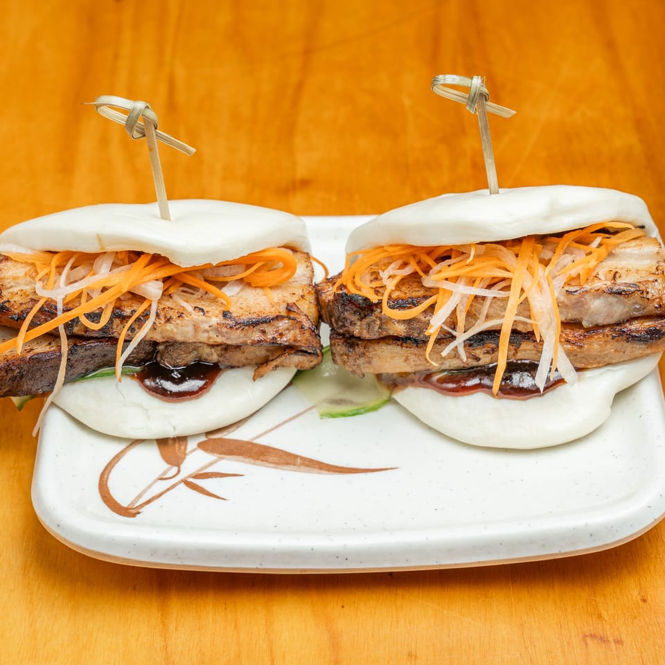 Two grilled pork bao sandwiches topped with shredded carrots and daikon on a decorative plate.