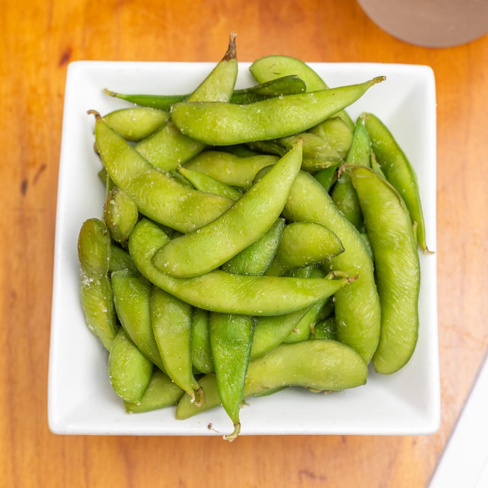 A white square dish holds steamed green edamame pods on a wooden table.