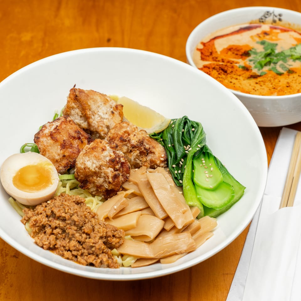 Ramen bowl topped with fried chicken, minced meat, bamboo shoots, greens, bok choy, and soft-boiled egg, accompanied by a creamy side dish.