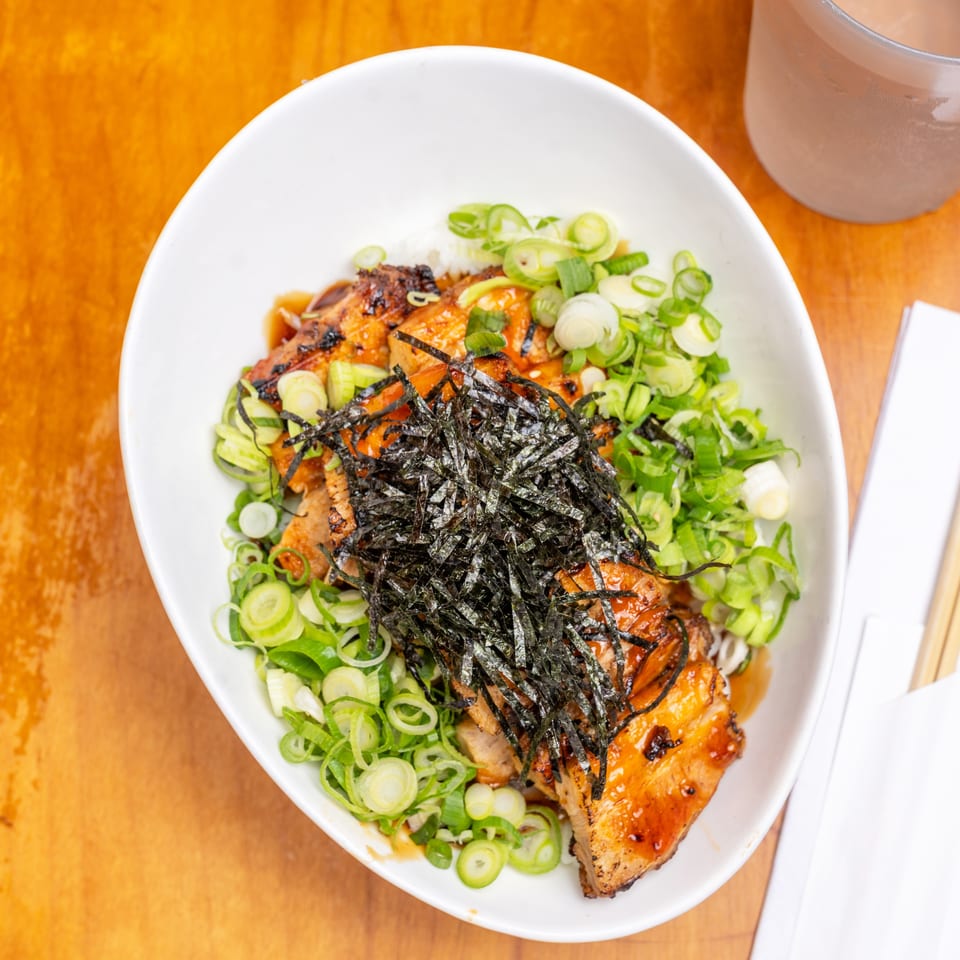 Japanese rice bowl topped with grilled salmon, sliced green onions, shredded nori, and sauce on a wooden table with chopsticks.