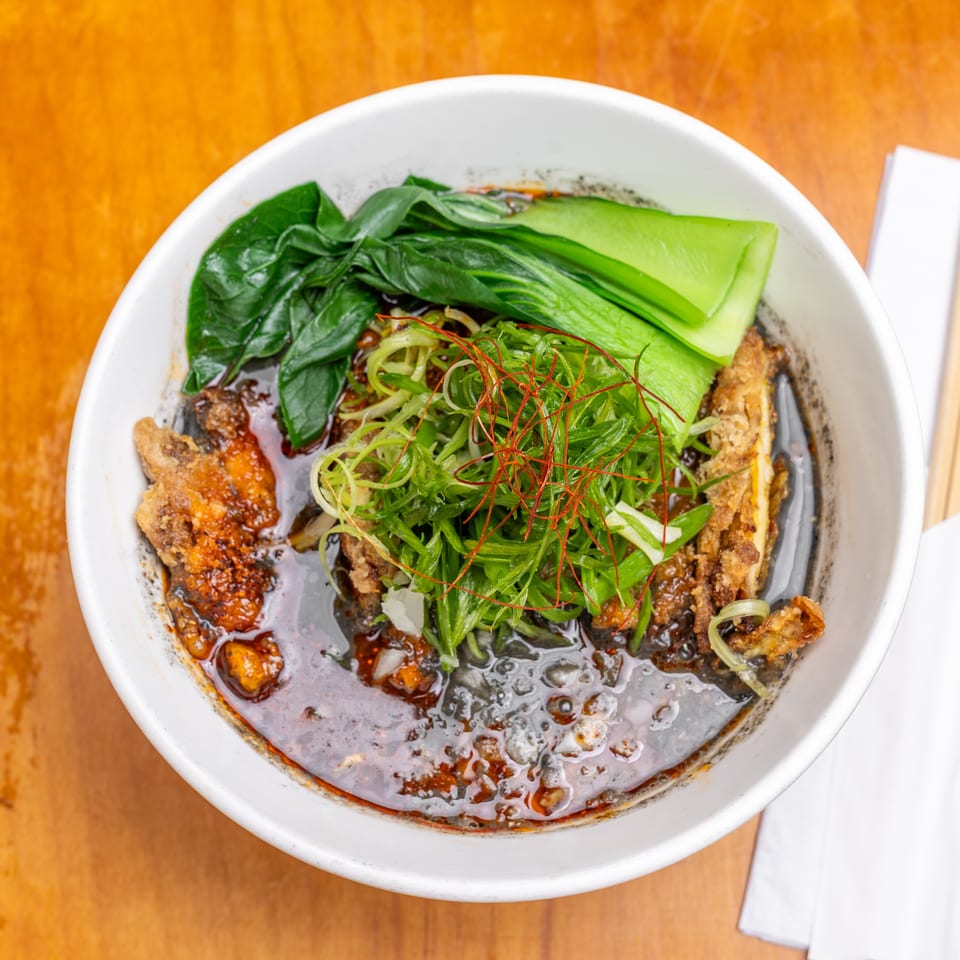 Top-down view of a bowl of spicy broth with fried fish, leafy greens, bok choy, scallions, and red chili threads.