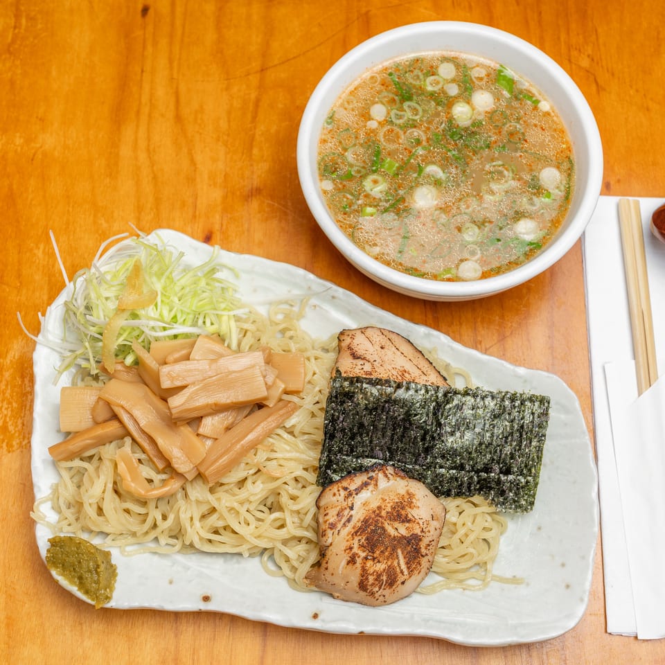 Overhead view of chilled ramen noodles topped with bamboo shoots, seaweed, grilled meat, and greens beside a bowl of scallion broth.