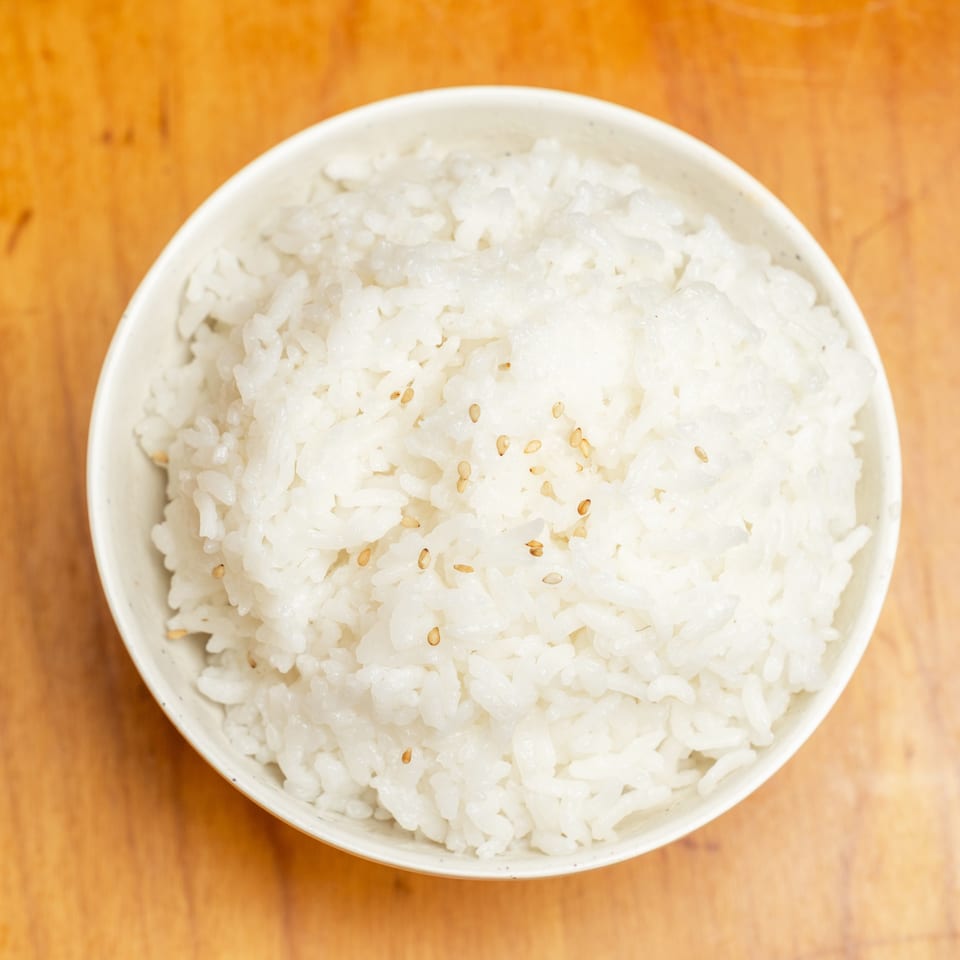 Overhead view of a bowl of steamed white rice sprinkled with sesame seeds on a wooden table.