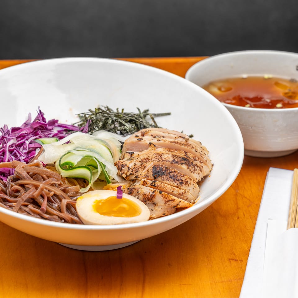 Ramen bowl topped with grilled chicken, soft-boiled egg, seaweed, purple cabbage, cucumber, and noodles beside dipping sauce.