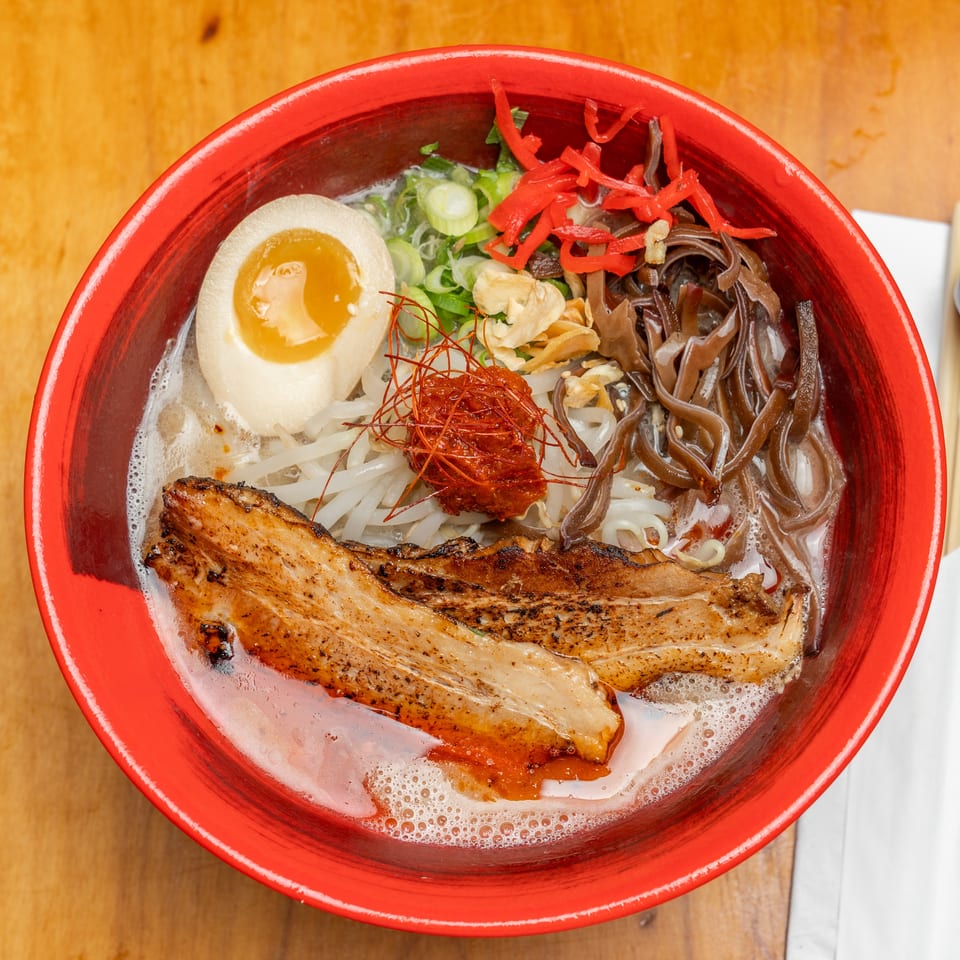Top-down view of a red bowl of ramen with grilled pork, soft-boiled egg, noodles, mushrooms, scallions, and chili garnish.