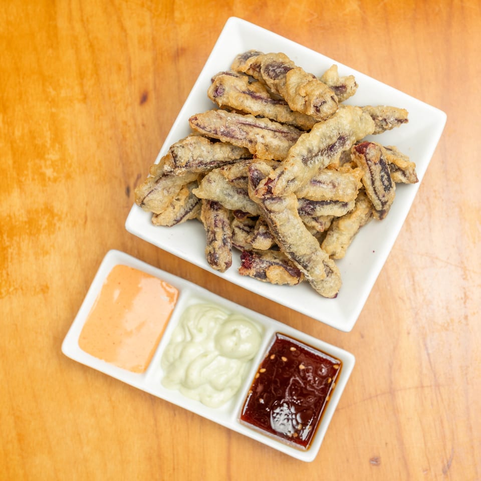 Crispy battered vegetable strips on a white plate with orange, green, and red dipping sauces on a wooden table.