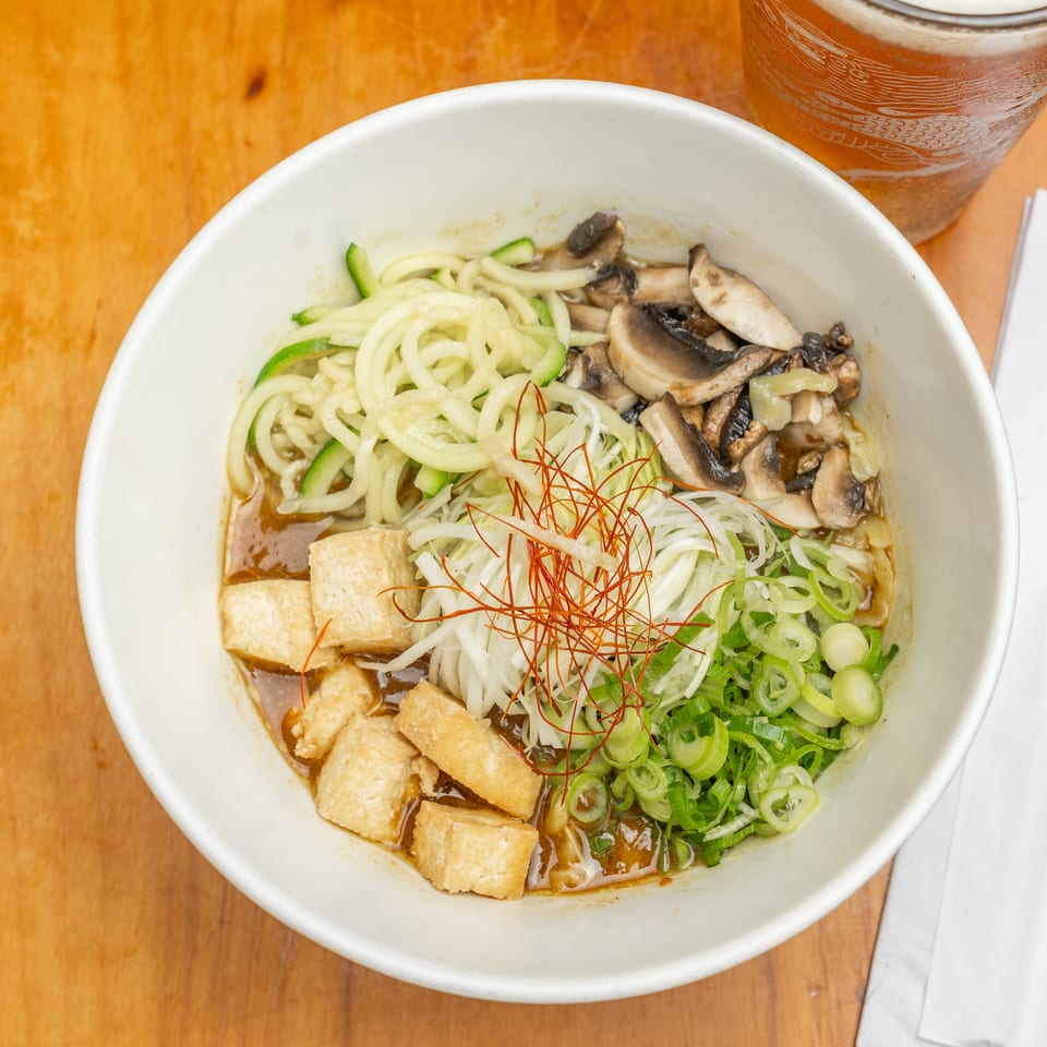 Overhead view of a Japanese noodle bowl with tofu, mushrooms, zucchini, scallions, chili threads, chopsticks, and iced tea.