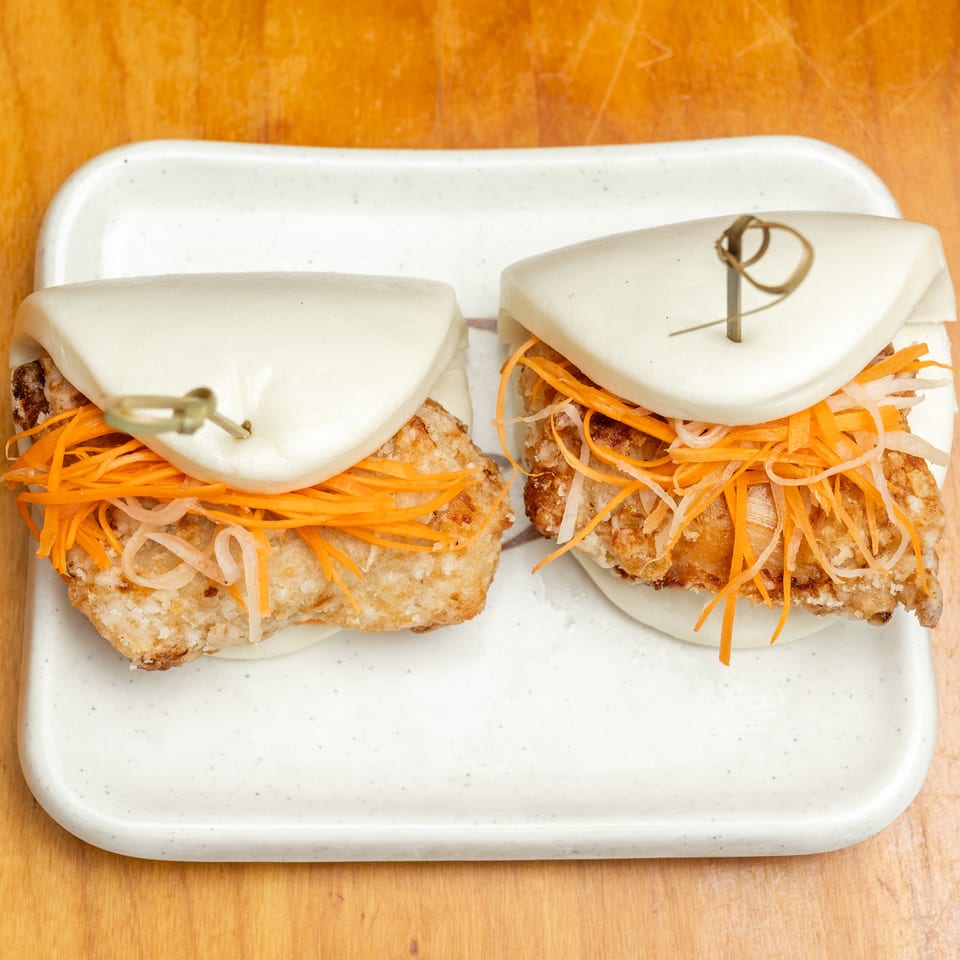 Two crispy chicken bao sandwiches with shredded carrots and daikon on a white rectangular plate.